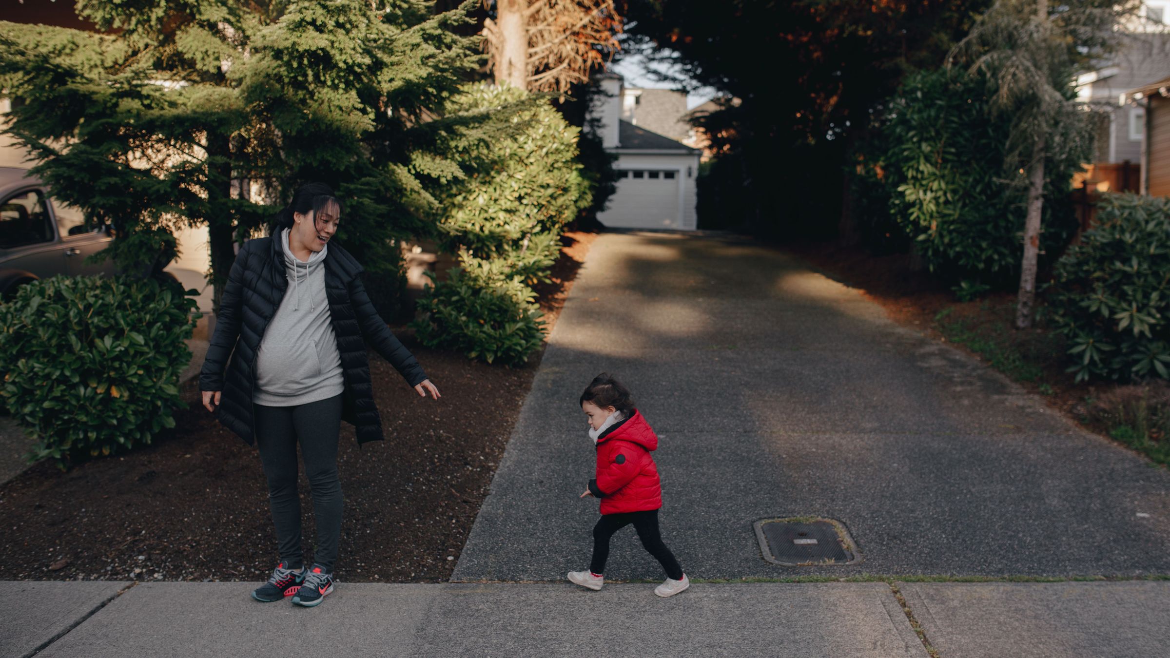 A smiling woman watches a toddler in a red jacket walk along a sunlit neighborhood driveway
